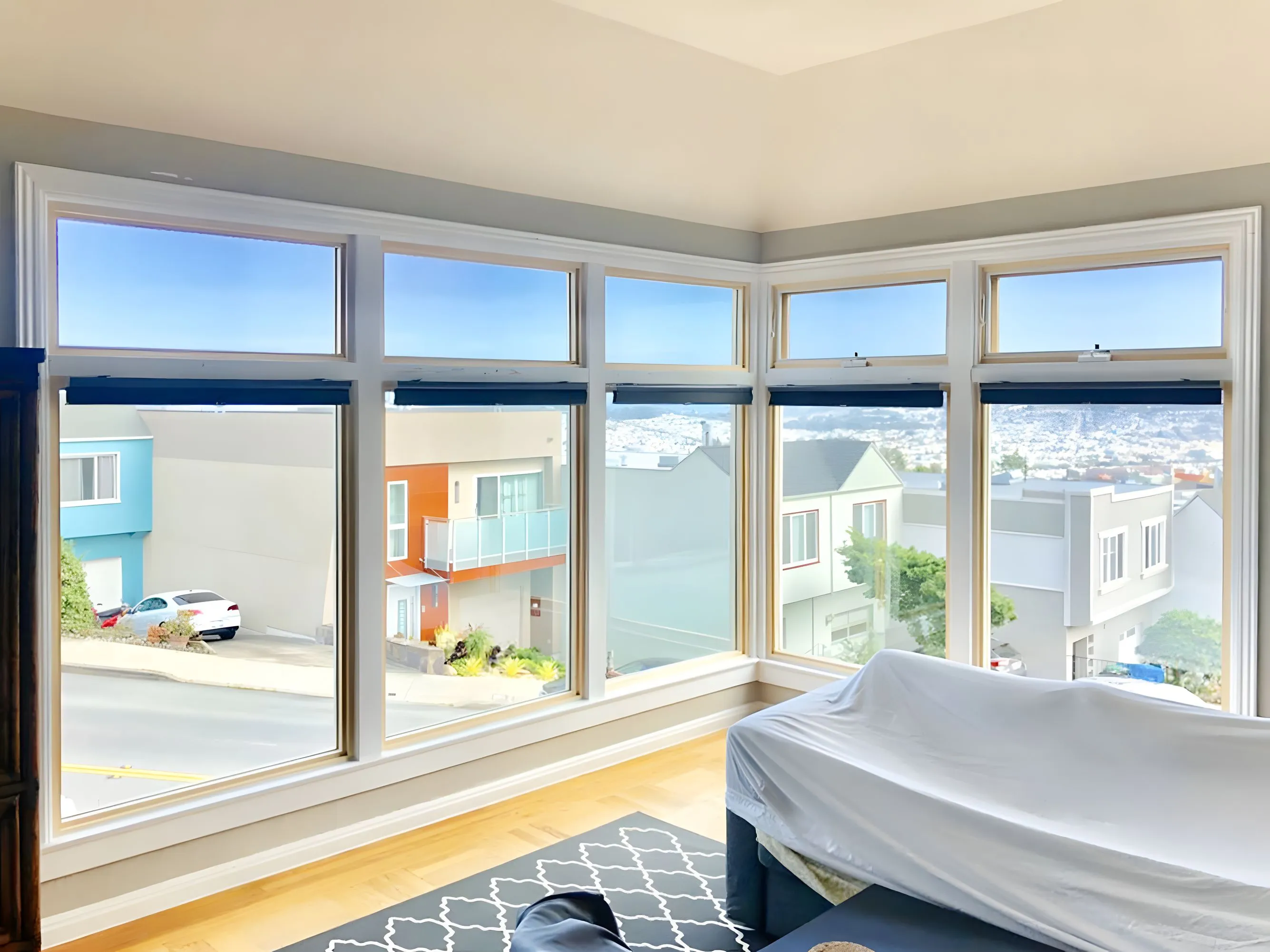Large bay windows in a modern living room, offering a clear view of colorful houses and a street outside. A covered sofa and patterned rug are partially visible. Large bay windows in a modern living room, offering a clear view of colorful houses and a street outside. A covered sofa and patterned rug are partially visible.
