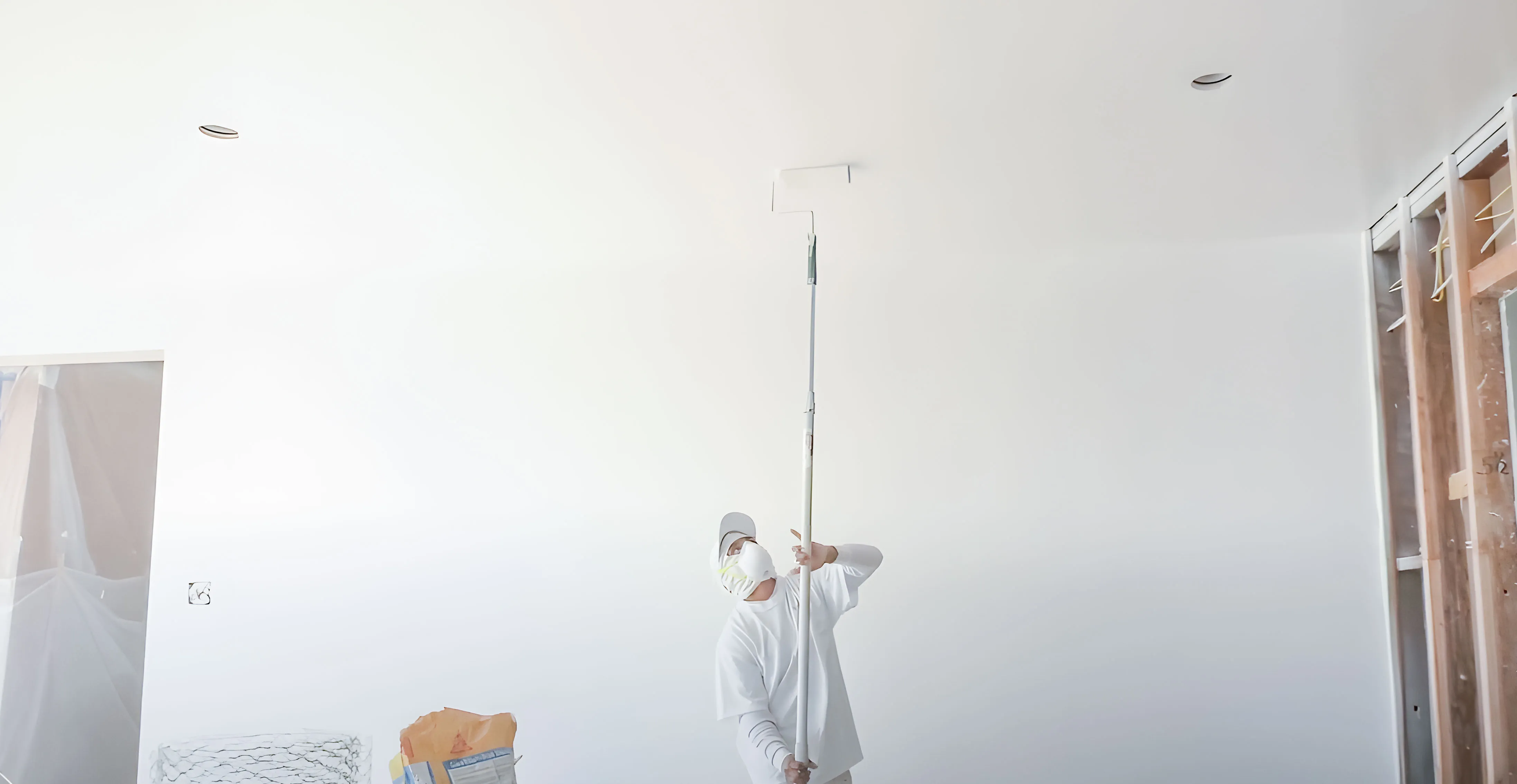 Person using a roller on a long handle to paint a ceiling white, wearing a mask and cap, with construction materials visible nearby. Person using a roller on a long handle to paint a ceiling white, wearing a mask and cap, with construction materials visible nearby.