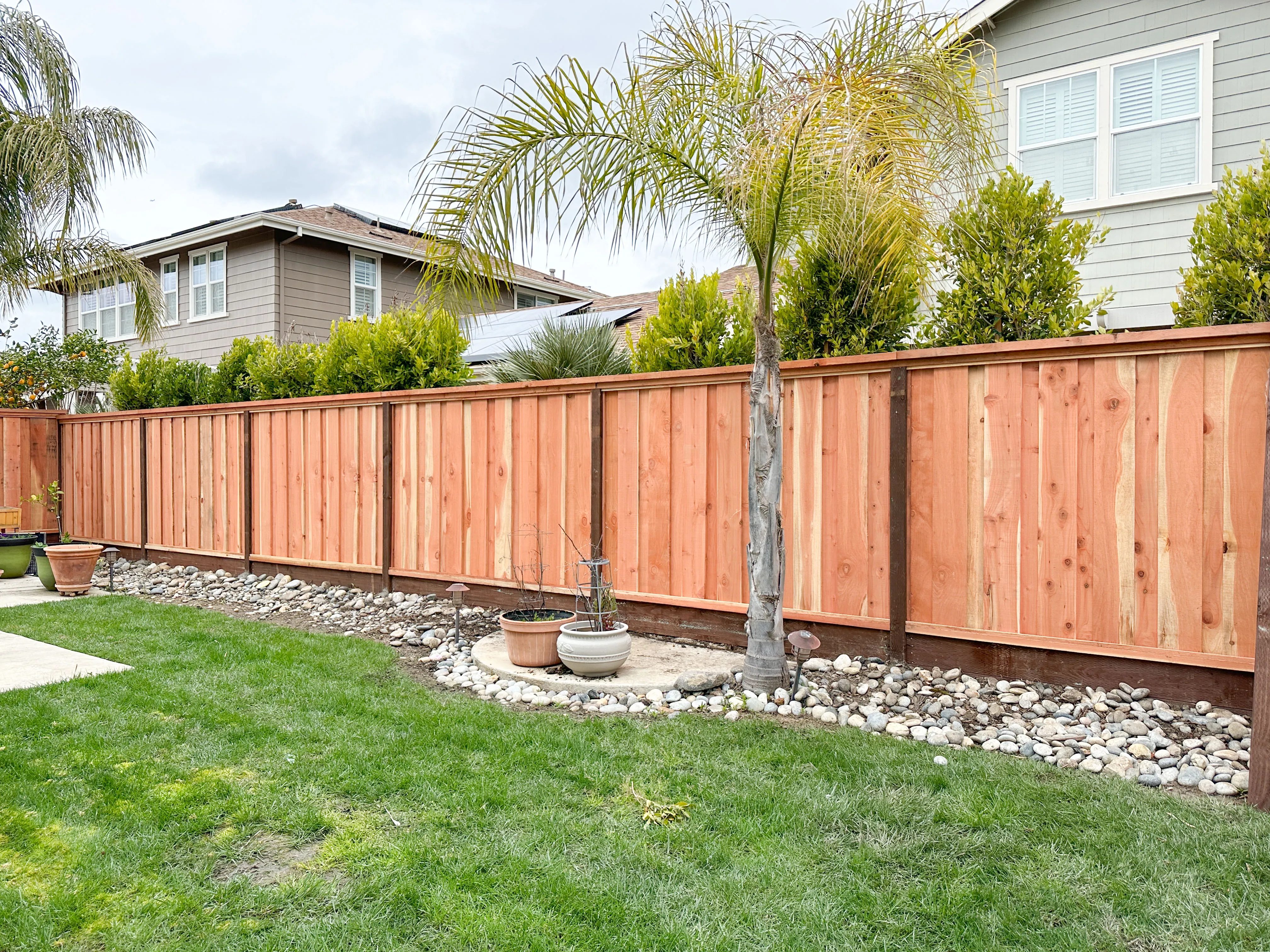 New wooden fence with natural finish along a landscaped yard, featuring green grass, potted plants, and a palm tree. New wooden fence with natural finish along a landscaped yard, featuring green grass, potted plants, and a palm tree.