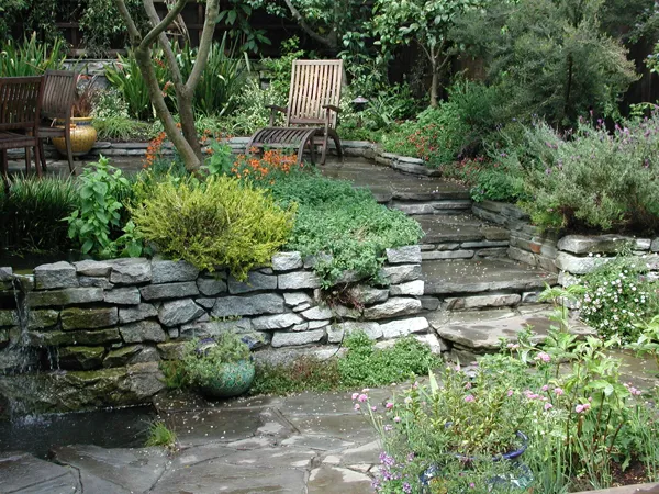Stone steps surrounded by various plants and flowers, with wooden chairs and a small water feature in a garden setting. Stone steps surrounded by various plants and flowers, with wooden chairs and a small water feature in a garden setting.