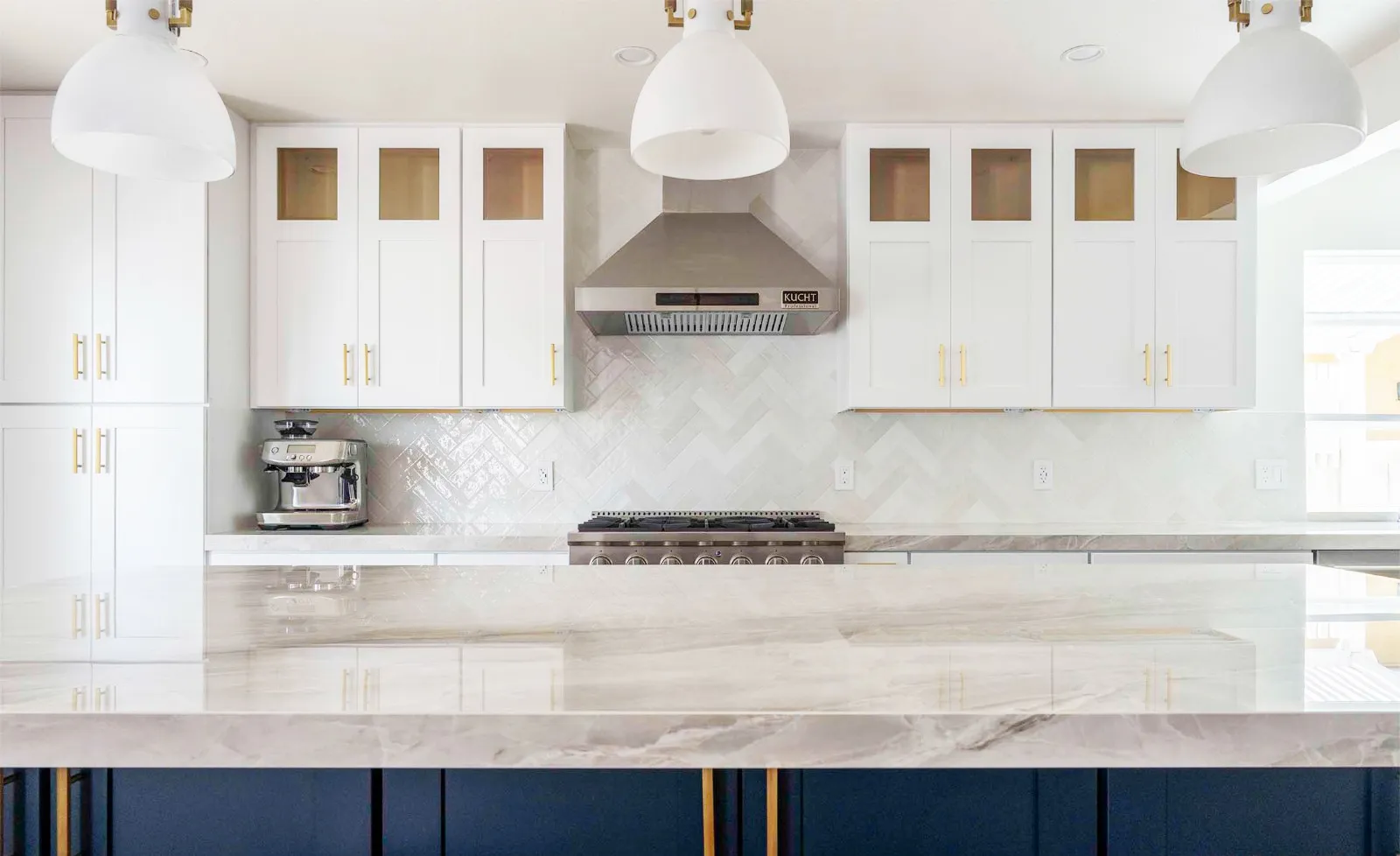 Modern kitchen with white cabinets, marble countertop, stainless steel range hood, and a coffee maker. Bright pendant lights hang above the island. Modern kitchen with white cabinets, marble countertop, stainless steel range hood, and a coffee maker. Bright pendant lights hang above the island.