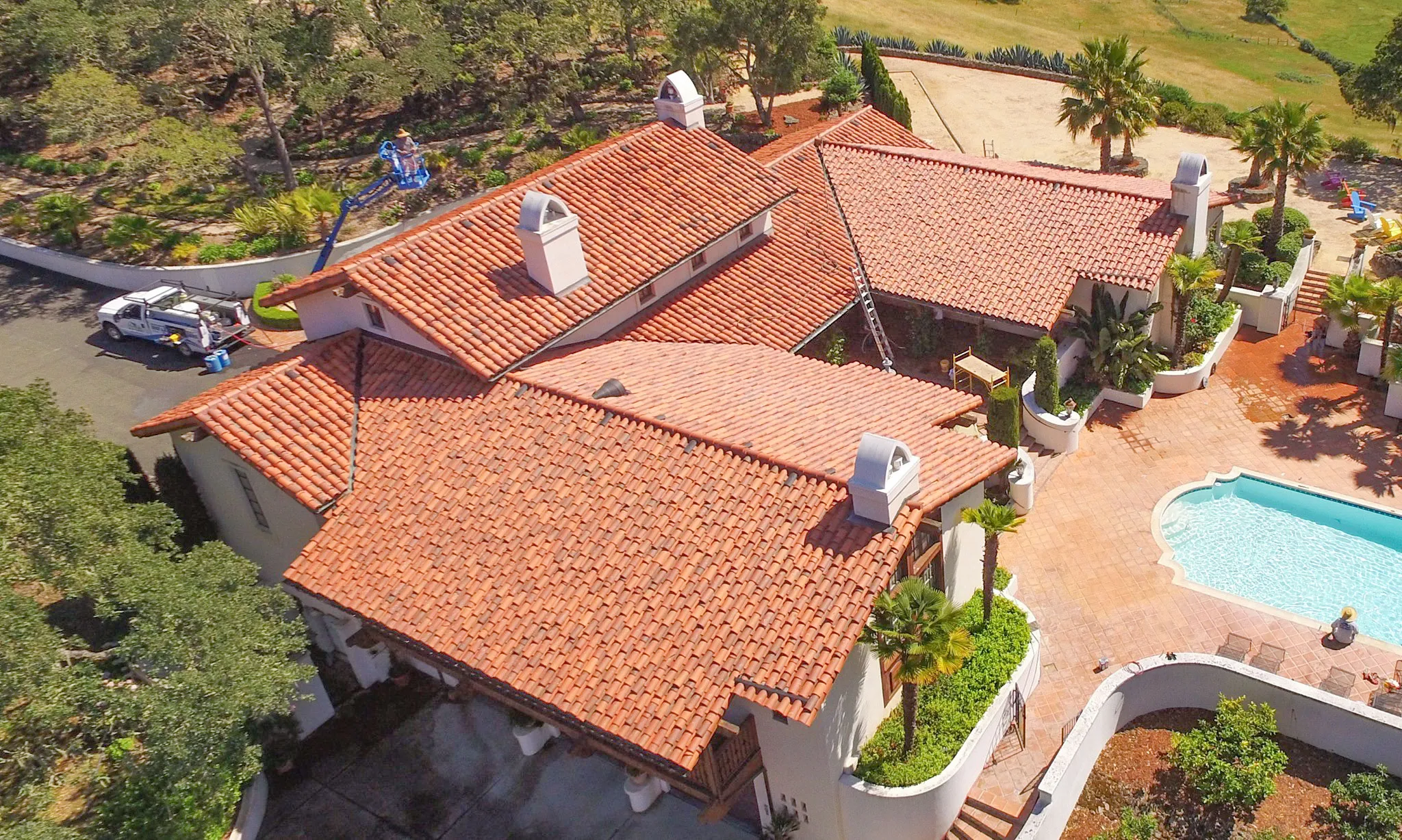  Aerial view of a Spanish-style home with a red tile roof, a lift truck, and workers attending to the roof. Surrounding landscape features trees and a pool.