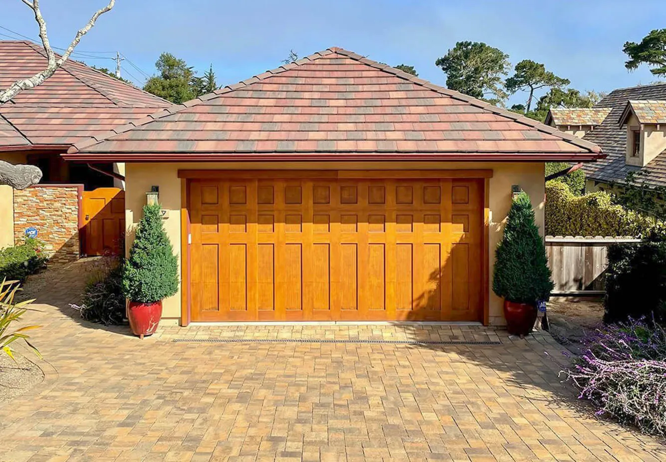  Garage with wooden door, framed by a brick driveway and roof tiles, flanked by potted plants.