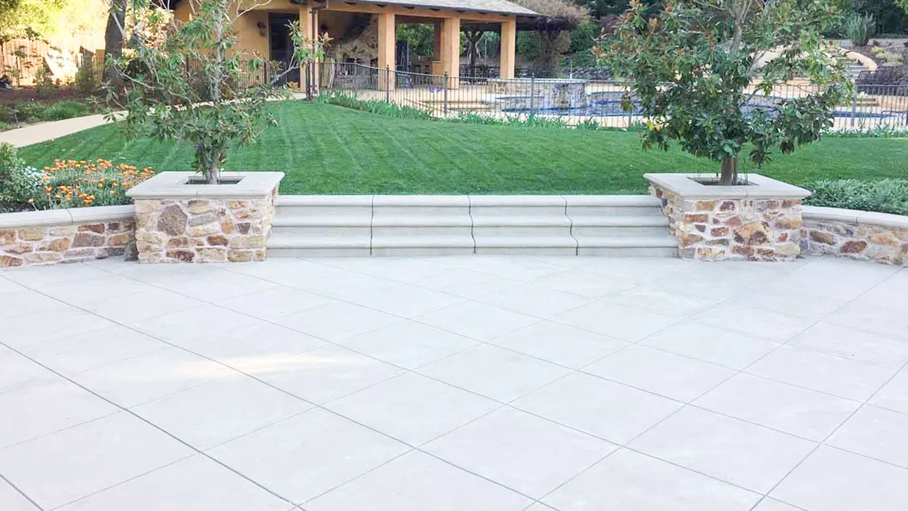  Spacious tile patio leading to grassy area with stone planters and trees, in front of a covered wooden gazebo.