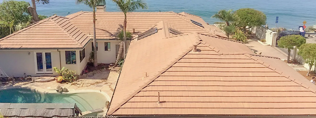  Aerial view of a house with a tiled roof, surrounded by trees, a pool, and ocean in the background.