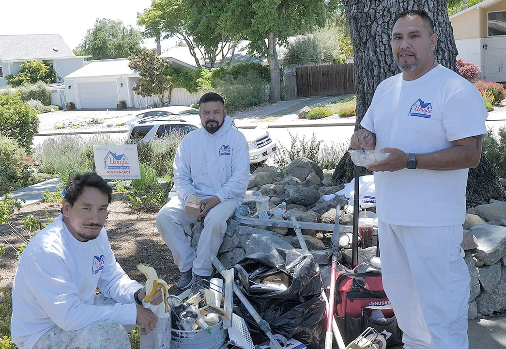 Three workers in white uniforms sit and stand around painting tools and supplies in a garden setting, taking a break.