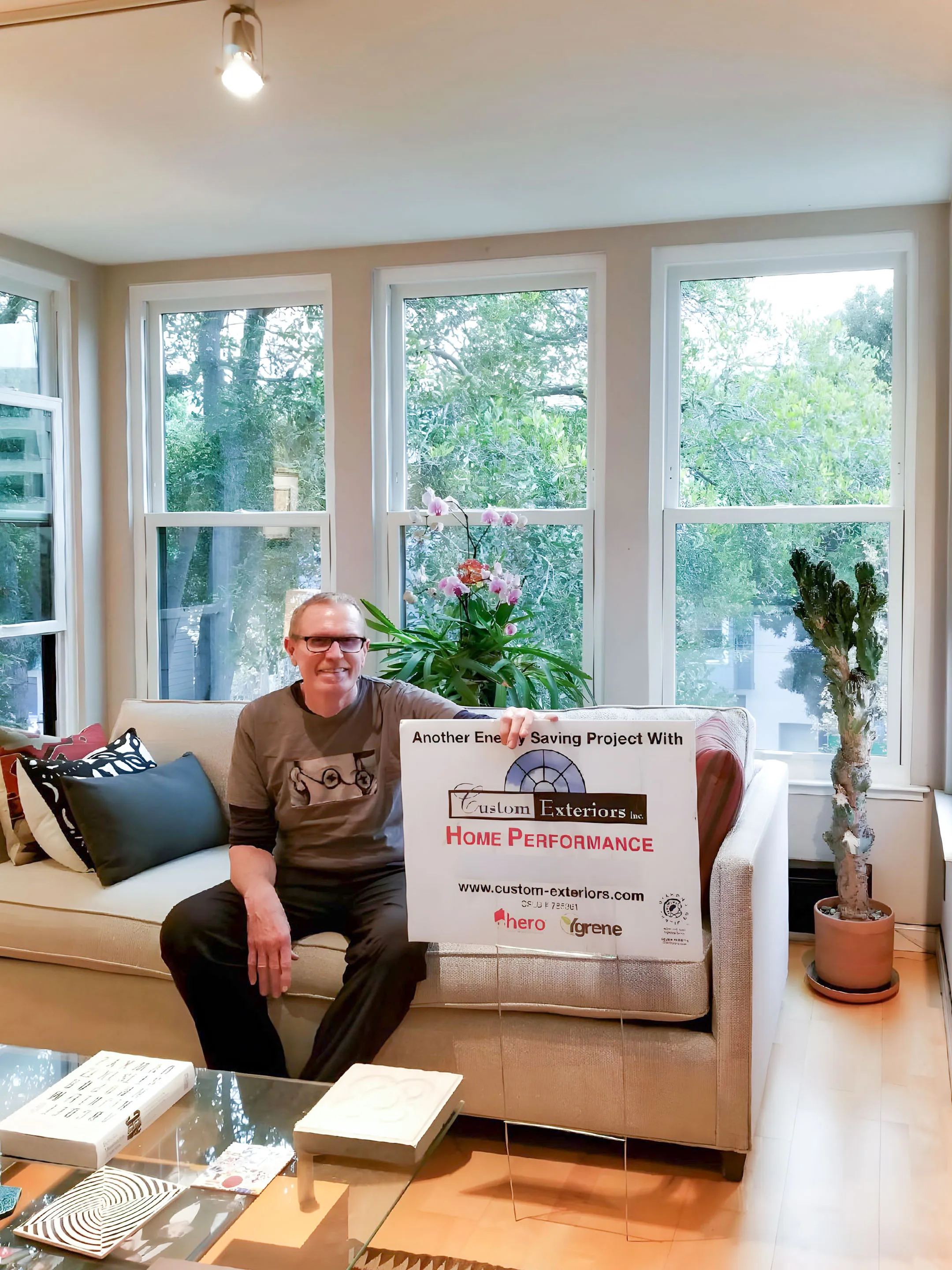 Picture of In a serene living room, a satisfied customer of Custom Exteriors, Inc. holds a sign highlighting their energy-saving project. Custom Exteriors, recognized for expertise and quality, transforms homes with precision. Copyright ©2025 Diamond Certified Resource - Custom Exteriors, Inc.