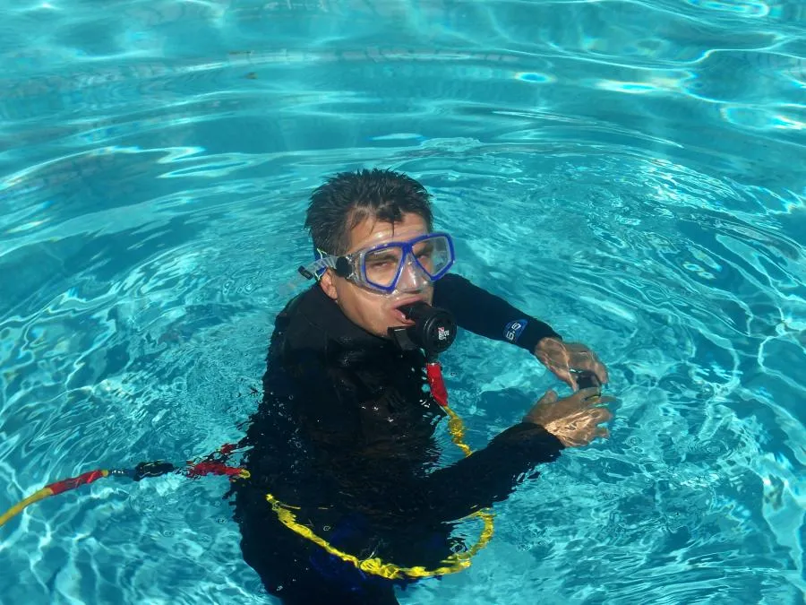 Diver in a pool wearing a snorkel mask and wetsuit, holding a tool, surrounded by clear blue water.