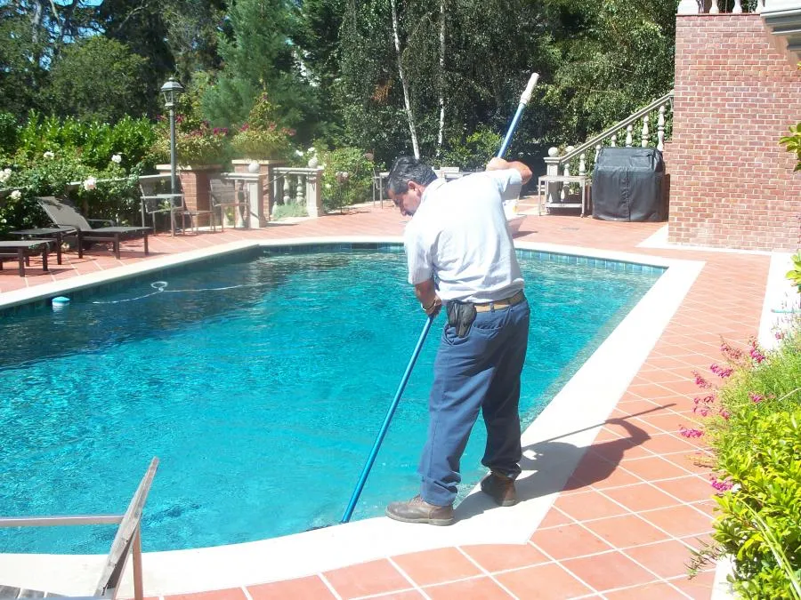  A technician uses a pool net to clean a residential swimming pool surrounded by a brick patio and lush greenery.