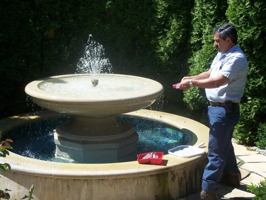 A technician works on a garden fountain, holding a water testing kit, with tools on the fountain edge and tall greenery in the background.
