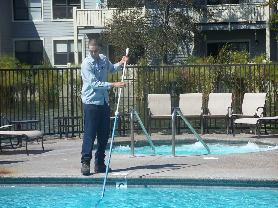  Technician using a pool skimmer to clean a swimming pool next to a hot tub, with lounge chairs and a building in the background.