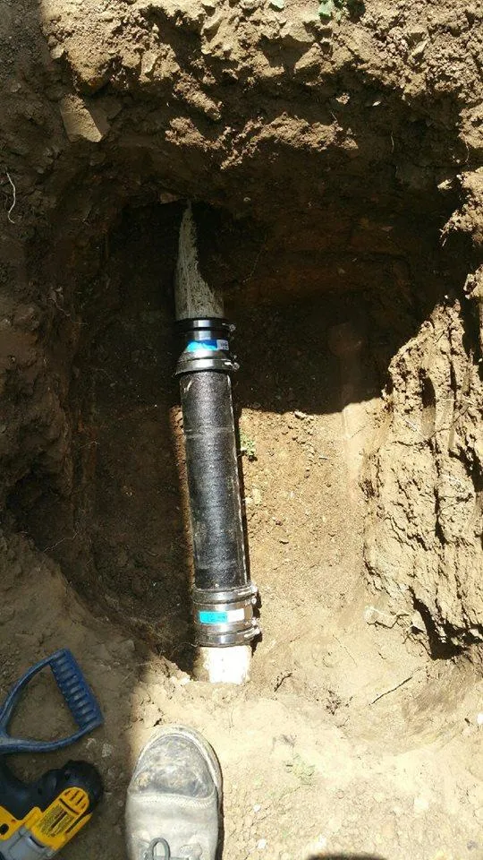 A newly installed black pipe with metal connectors in a dirt trench, with a shoe and a tool nearby.