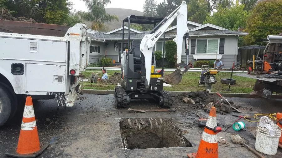  A small excavator digs a hole in a suburban street, surrounded by traffic cones, trucks, and work tools, with workers in the background.