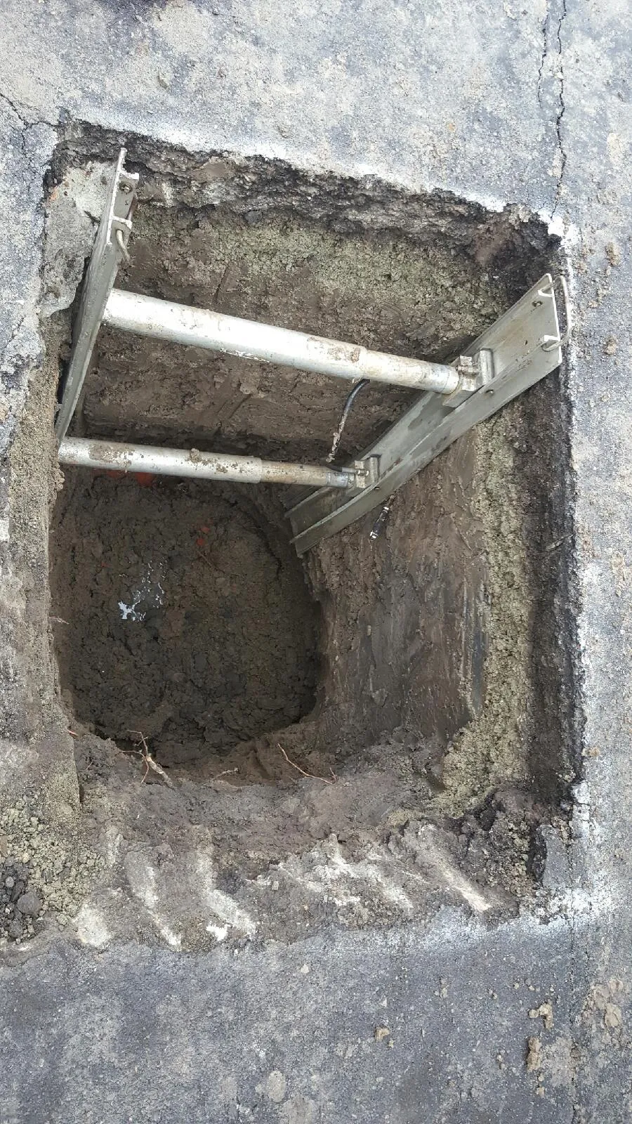  A square hole dug into a concrete surface, revealing soil and a metal bracket, indicating a plumbing access point.