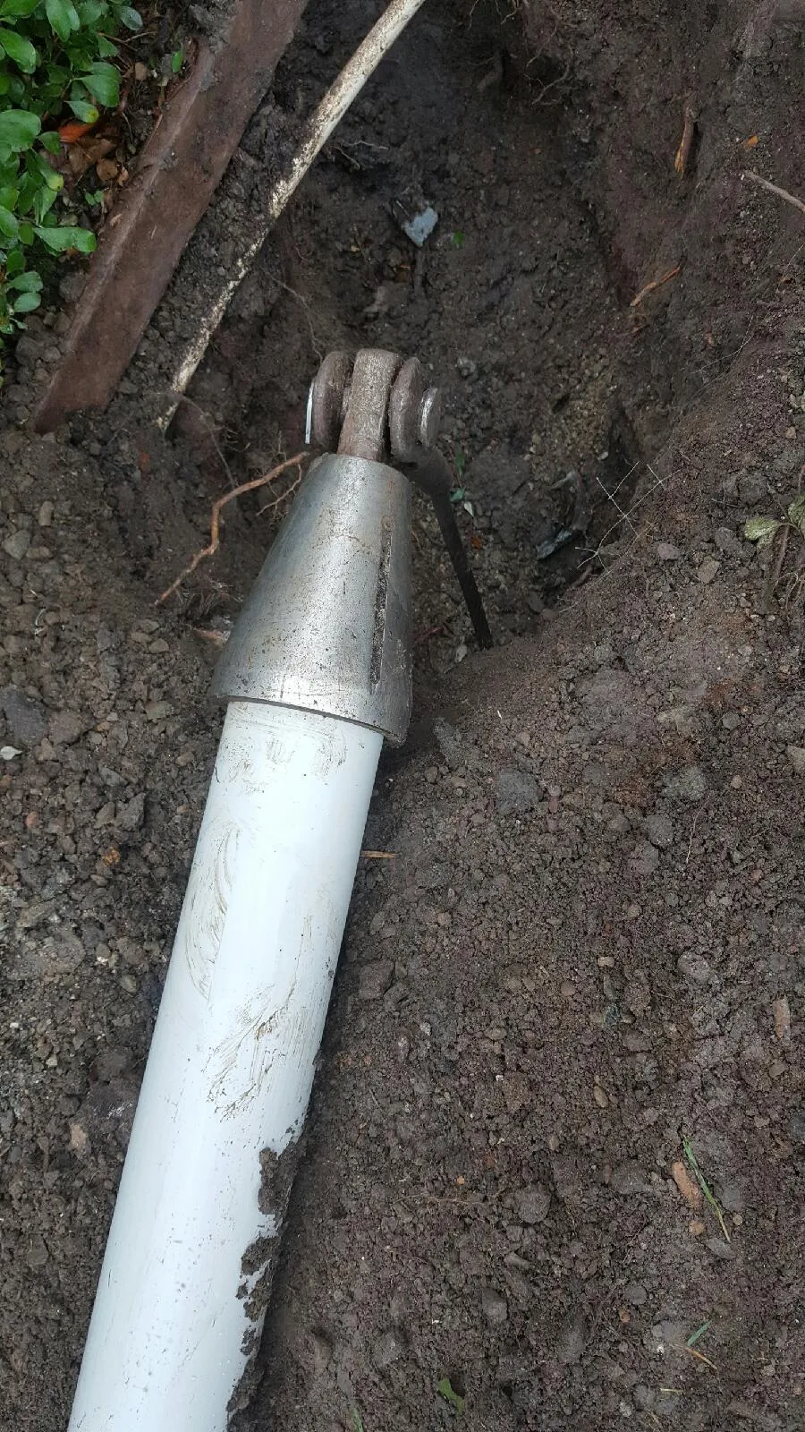  A close-up of a metal and plastic pipe joint exposed in a trench, surrounded by soil and some greenery.