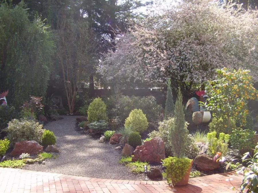  A garden path winds through a variety of green shrubs, flowering plants, and trees, surrounded by rocks and a brick patio.
