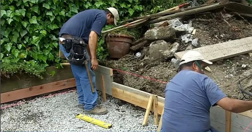  Two workers in blue shirts and caps adjust a wooden frame on a gravel surface, with a green leafy background and construction tools nearby.