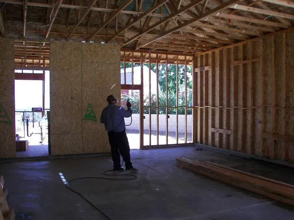  Worker sprays chemicals on wooden frame inside an unfinished structure, surrounded by construction materials.
