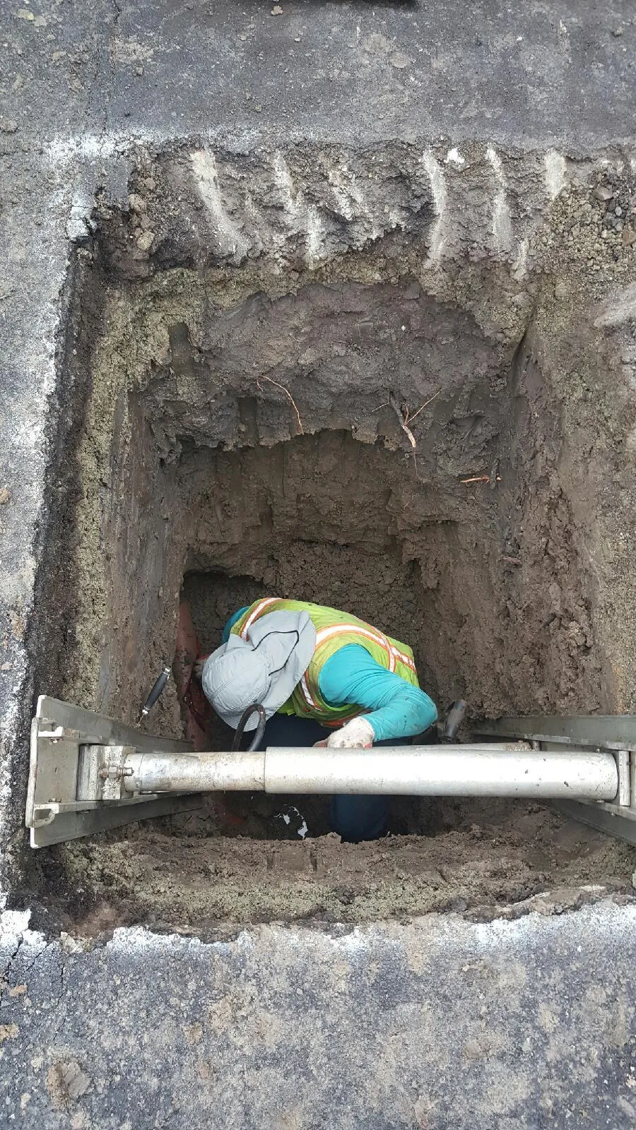  Worker in a trench repairing underground pipes, wearing a safety vest and hat, surrounded by dirt and road asphalt.