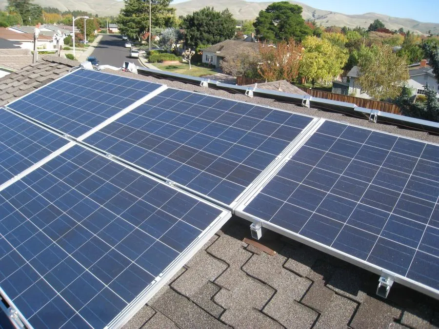  Solar panels are installed on a shingled rooftop in a suburban neighborhood, surrounded by trees and distant hills.