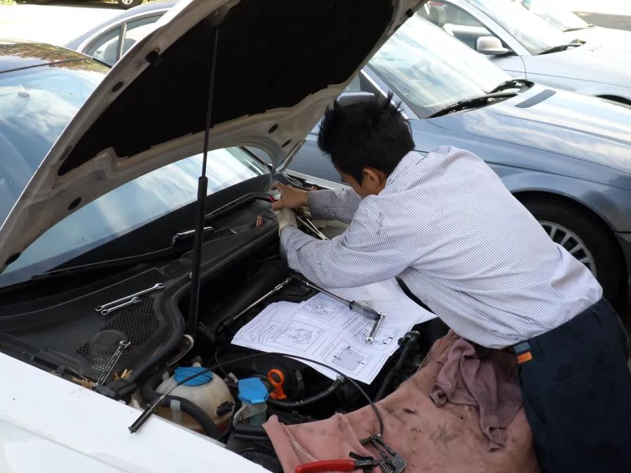  A mechanic repairs a car engine using tools and manuals under the open hood in a parking lot.