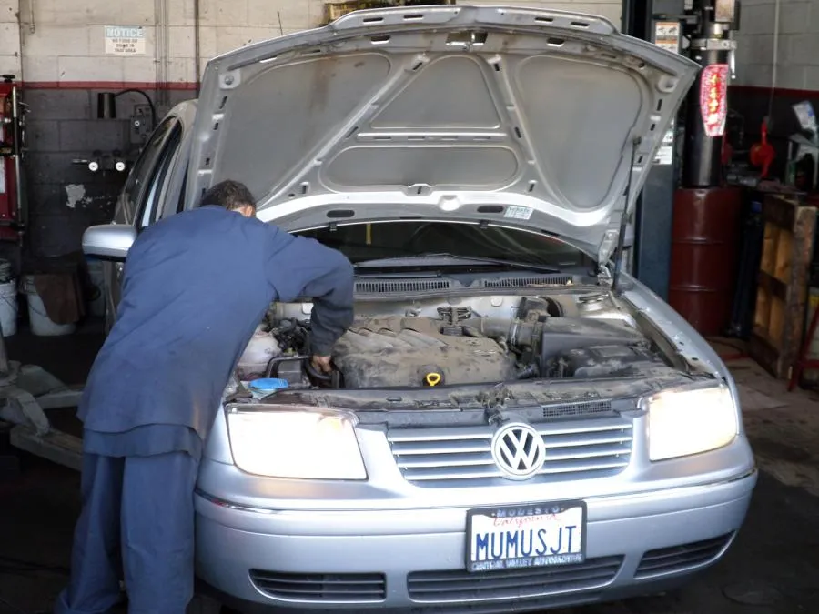  Mechanic in blue overalls inspects the engine of a silver Volkswagen with the hood open inside an auto repair shop.