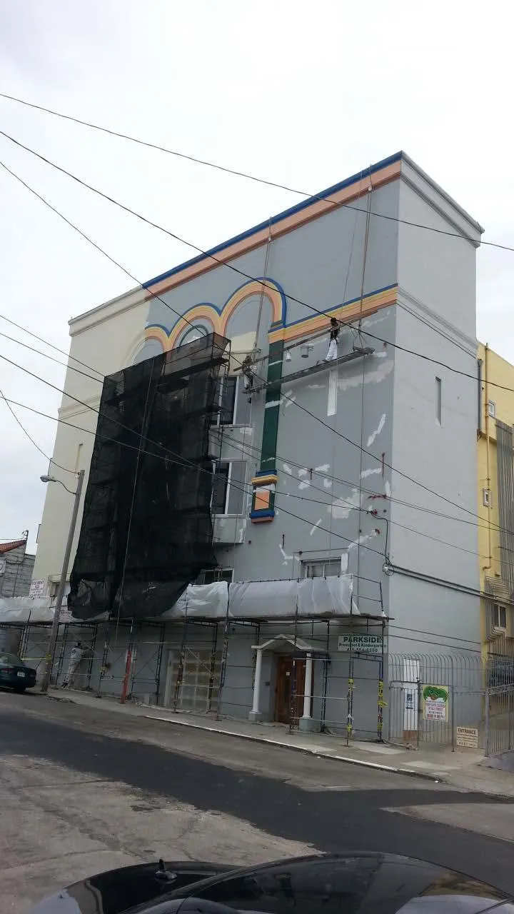  A painter works on a building facade, adding colorful arches. Scaffolding and netting are visible; street and cars are in the foreground.