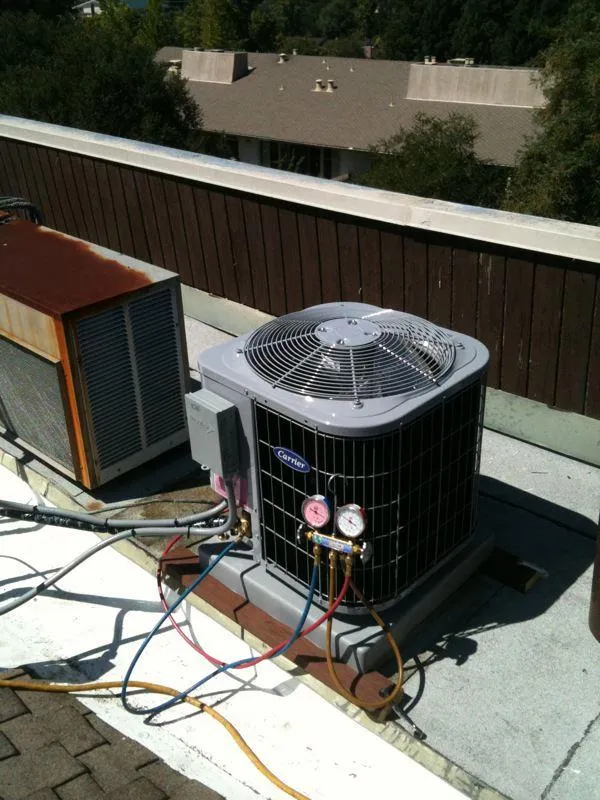  Rooftop HVAC unit with gauges attached, surrounded by cables, next to another old unit, set against a backdrop of trees and buildings.
