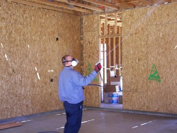 A person wearing a mask and gloves sprays treatment inside a building under construction, with exposed wooden walls and ceiling beams.