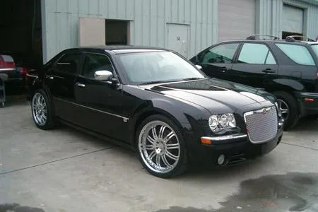  Black car with chrome rims parked in front of a garage, adjacent to another vehicle.