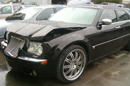 Black car with a damaged front end and large wheels parked outdoors on a wet surface.