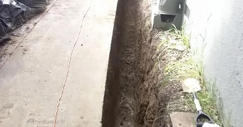  A narrow trench runs beside a building with a shovel and some grass visible, indicating construction work.