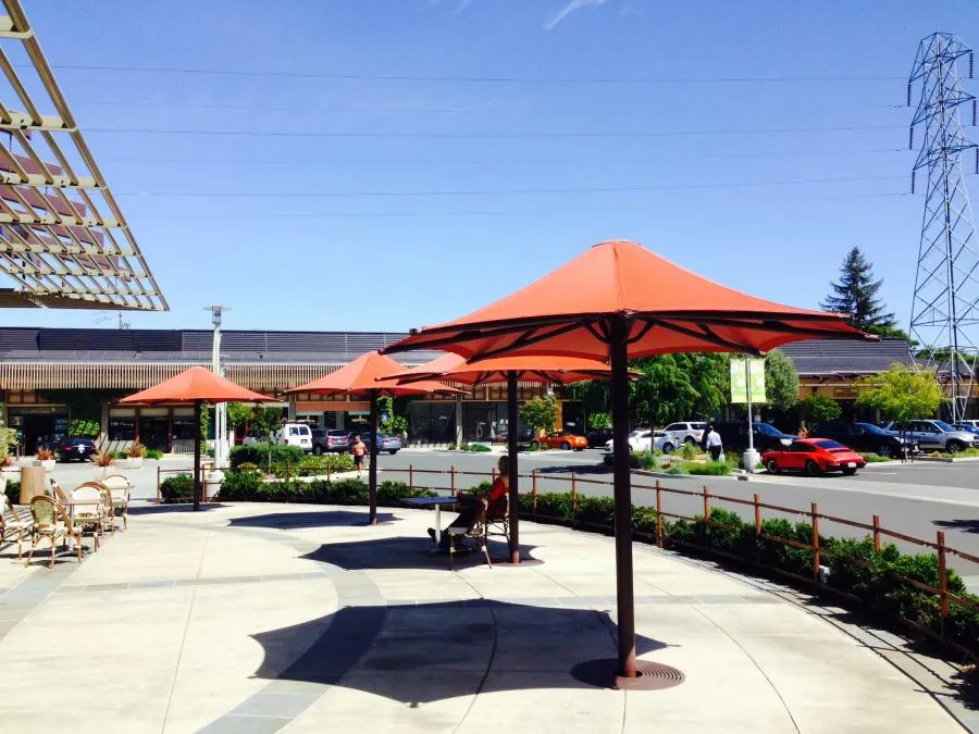 Outdoor area with orange sunshades, chairs, and tables. Nearby parking lot with cars and a power line. Clear sky overhead.