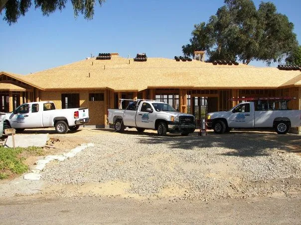 Three white trucks with company logos parked at a construction site in front of a partially constructed house with wooden framing.