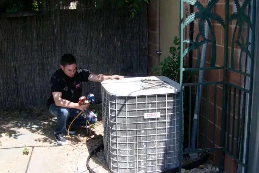  A technician checks an outdoor air conditioning unit with tools, surrounded by plants and a metal trellis.