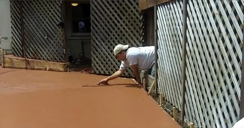  Person smoothing freshly poured concrete with a trowel next to lattice fencing.