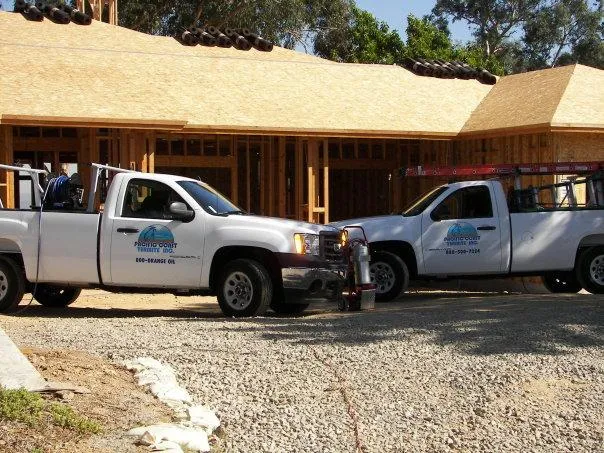 Two Pacific Coast Termite trucks parked in front of a wooden house under construction, with tools and a ladder visible. Gravel driveway in the foreground.