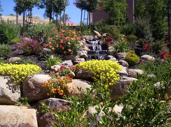  Flowing water over stacked stones surrounded by colorful flowers and lush greenery in a landscaped garden setting.
