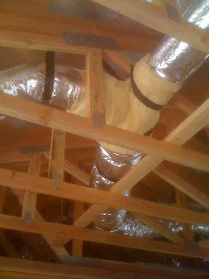  Ductwork in an attic with foil-covered insulation, secured by straps to wooden beams.