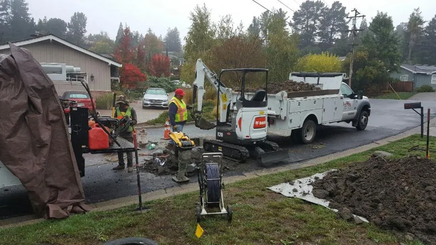  Workers in safety vests operate an excavator and other equipment on a residential street, with a truck and tools nearby.