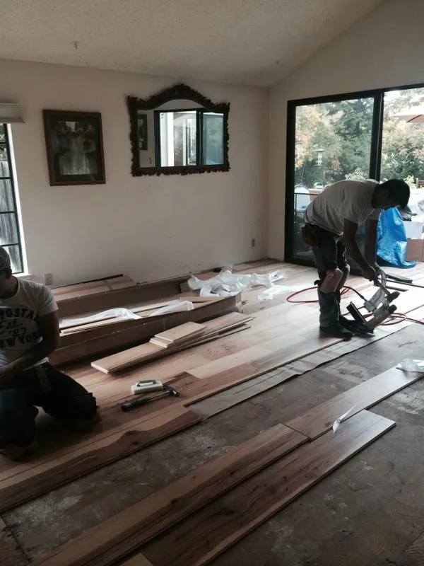  Two workers install hardwood flooring in a room with tools and materials scattered around. One kneels, while the other uses a flooring nailer.