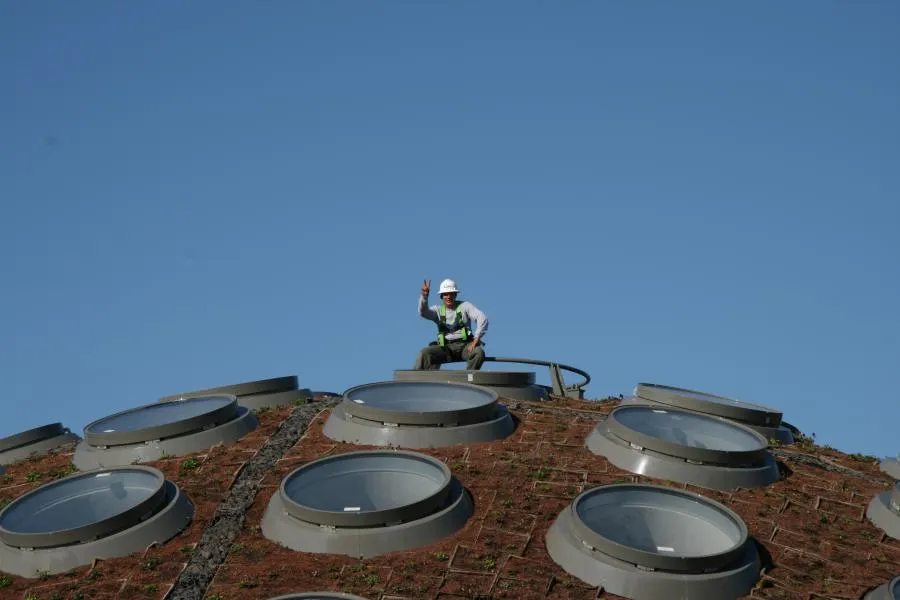  A worker in safety gear waves from a dome roof with multiple skylights against a clear blue sky.