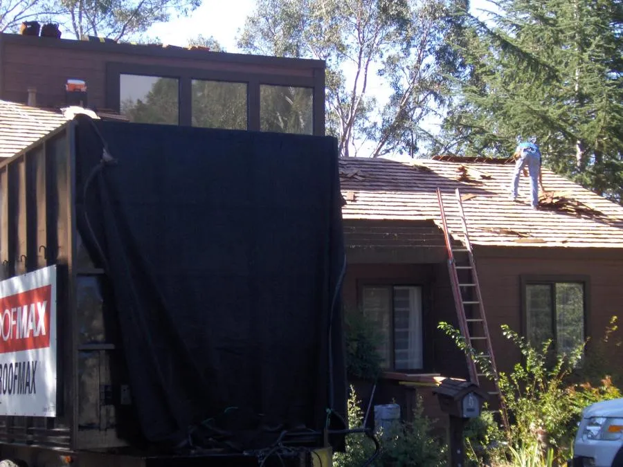 Worker on a roof, standing near a ladder, with roofing materials scattered. A truck with the Roofmax logo is parked nearby. Trees surround the house.