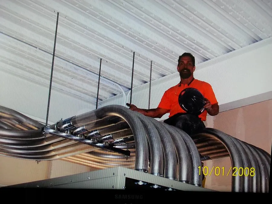  Technician in an orange shirt working on curved metal conduits near a ceiling.