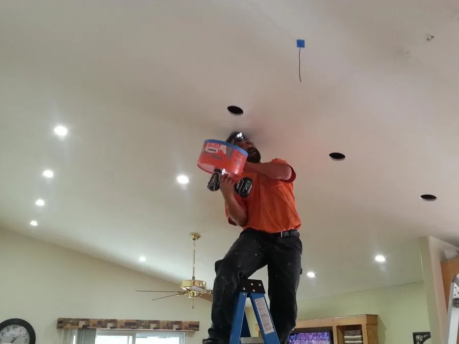  Electrician on ladder installing ceiling lights with a drill, surrounded by recessed lighting fixtures.