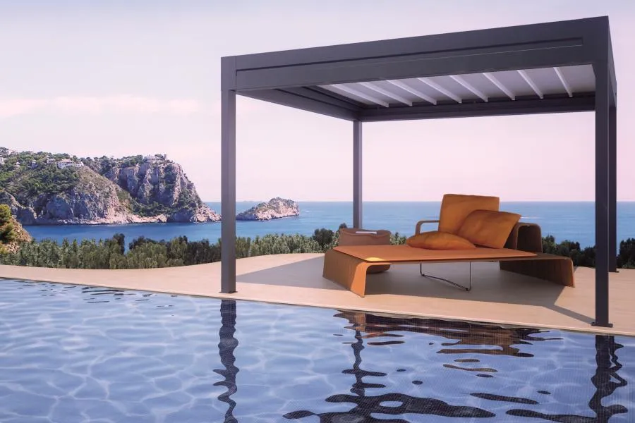A modern pergola with orange cushions overlooks an infinity pool and scenic coastline under a clear sky.