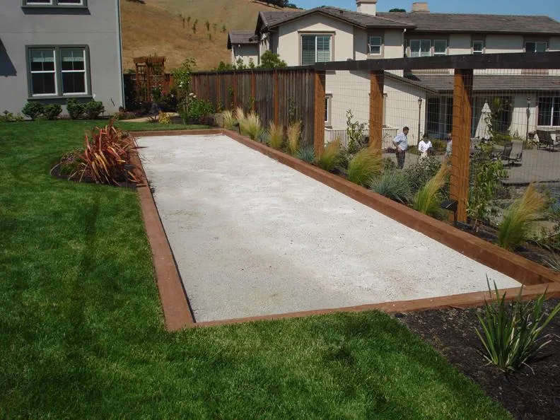  A bocce ball court in a landscaped backyard with grass, plants, and a wooden fence. Two people stand near a patio area.