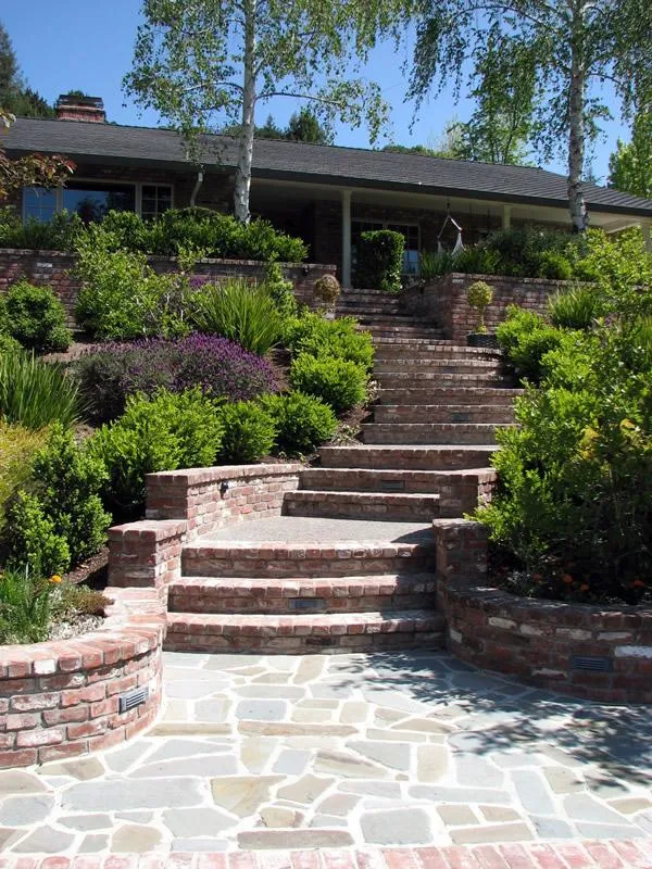  Brick steps and pathways surrounded by green shrubs and trees, leading up to a house with a porch.