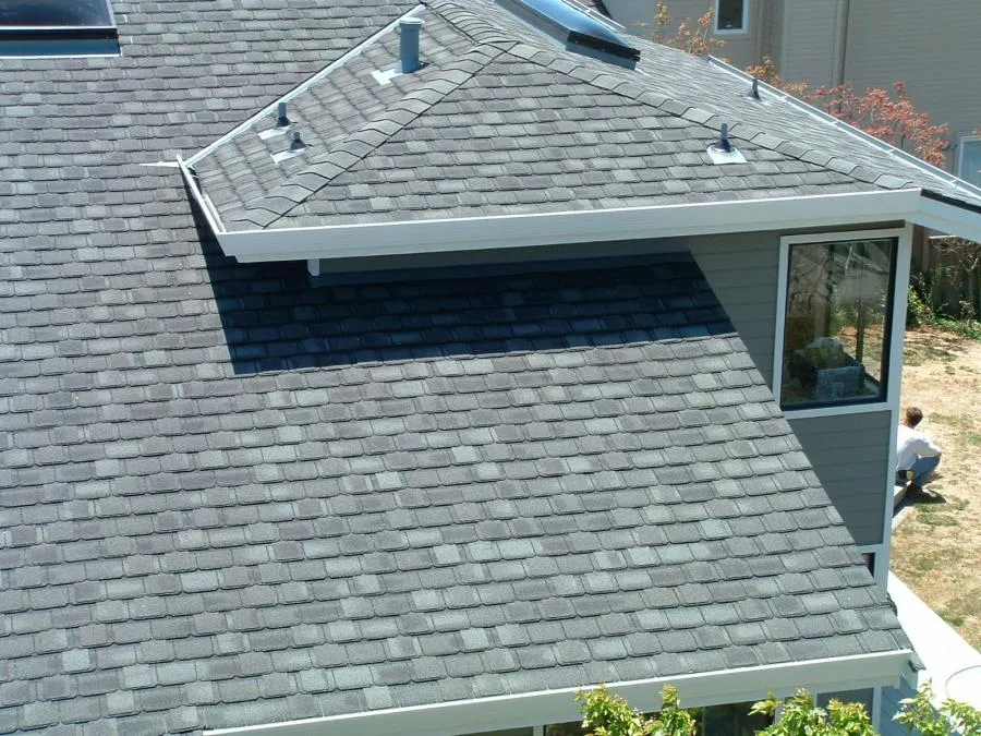  A well-finished shingle roof with skylight and vents, adjacent to a windowed wall, with a person working near the building.