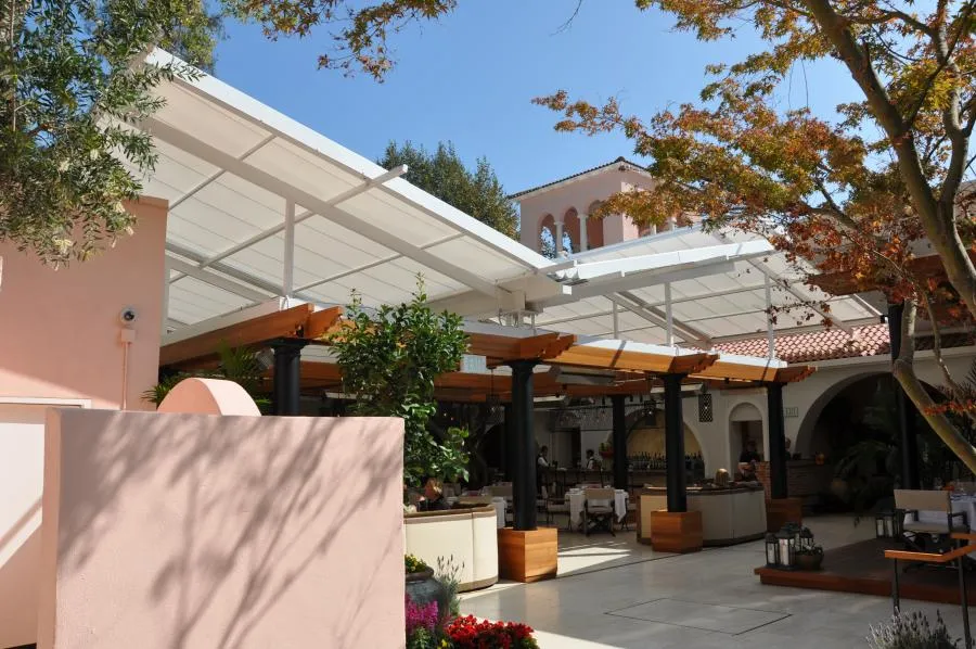 A bright courtyard with modern sunshades, tables, chairs, and surrounding trees. The sunshades are supported by black and wooden beams, providing a shaded area.
